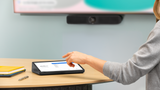 A photograph of a Logitech Tap on a table in a meeting space. A person’s hand is hovering over the Logitech Tap. In the background, there is a Logitech MeetUp 2 mounted below a TV screen.