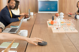 An image of the Rally Mic Pod in graphite mounted on a table in a meeting room. A hand from one of the meeting participants sat around the table is reaching over and pressing the mute button on the mic pod.