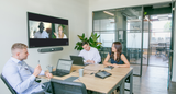 An image of a small meeting space with three participants sitting at a table. At the front of the room, there is a TV mounted on the wall showing an active Google Meet call. A Logitech Rally Bar Mini is mounted below the screen, and, on the table, a Logitech Tap is lit up with meeting controls.