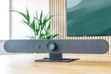An image of the Logitech Rally Bar Mini on a table in a meeting room. Behind the Rally Bar Mini, there is a TV screen mounted on the wall.