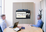 An image of a meeting space where two participants are sitting at a table facing towards a TV screen which is mounted to the wall. The screen is showing an active Google Meet call. A Logitech Rally Bar is mounted above the screen, and, on the table, a Logitech Tap is lit up with meeting controls.