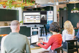 An image of an open meeting area where four participants are facing towards a TV screen showing an active Google Meet call. The screen is mounted on a cart along with the Logitech Rally Bar and the Logitech Tap.