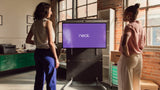 A photo of two women standing in front of the Neat Board 50 which is mounted on the Neat adaptive stand. On the screen is the Neat logo on a purple background. The background of the room shows large windows, brick walls and various office items, including a printer and cardboard boxes.